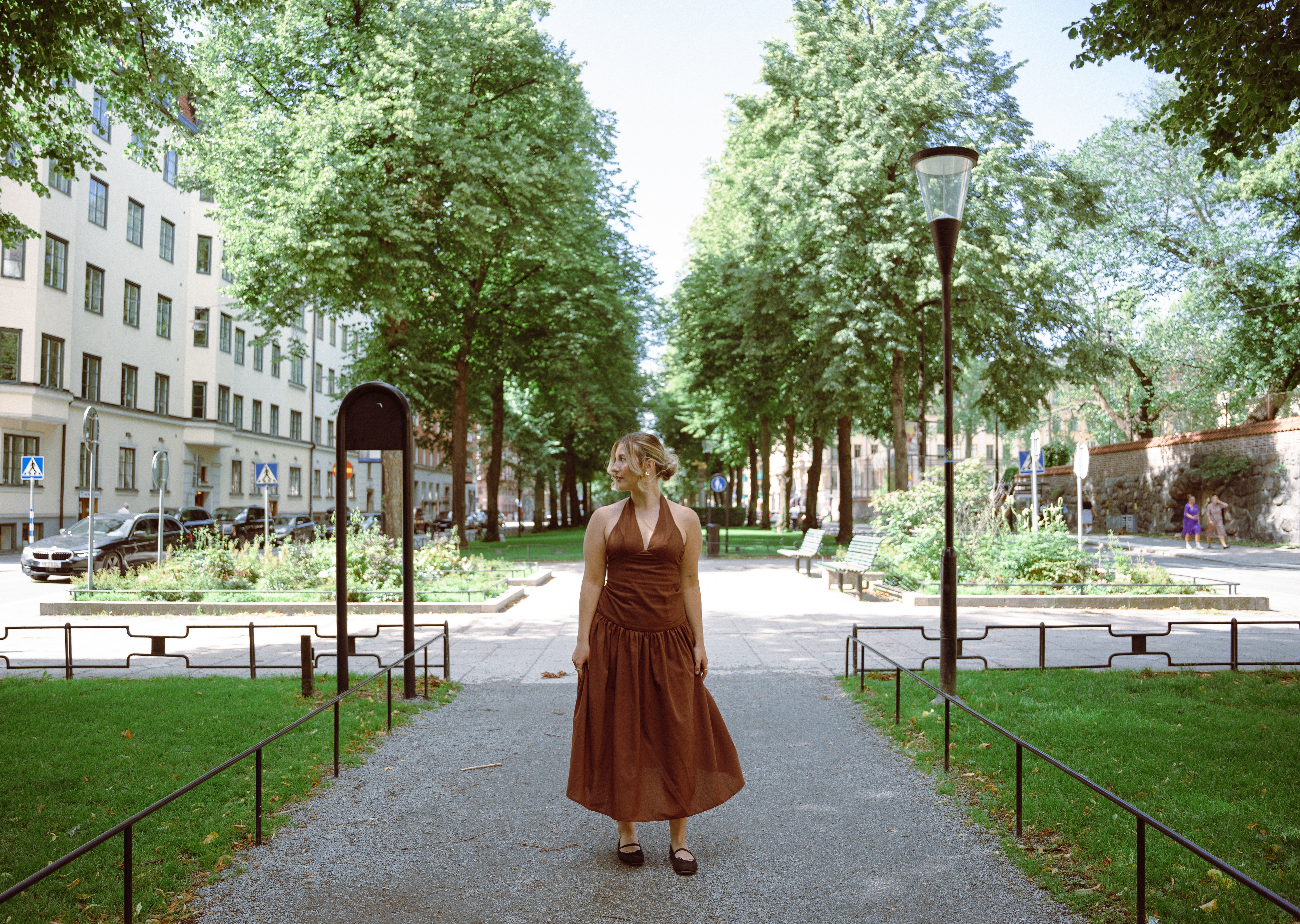 Woman in a brown dress standing on a tree-lined path in an urban park.