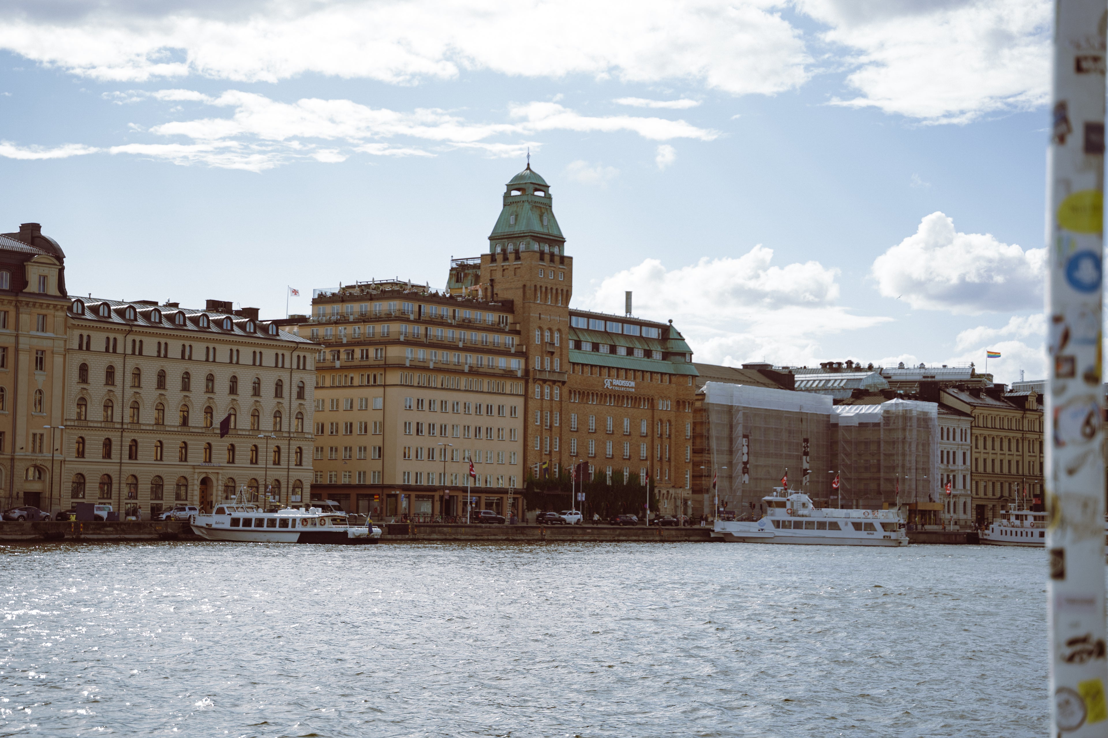 Cityscape with buildings and boats on a water body under a blue sky with clouds.
